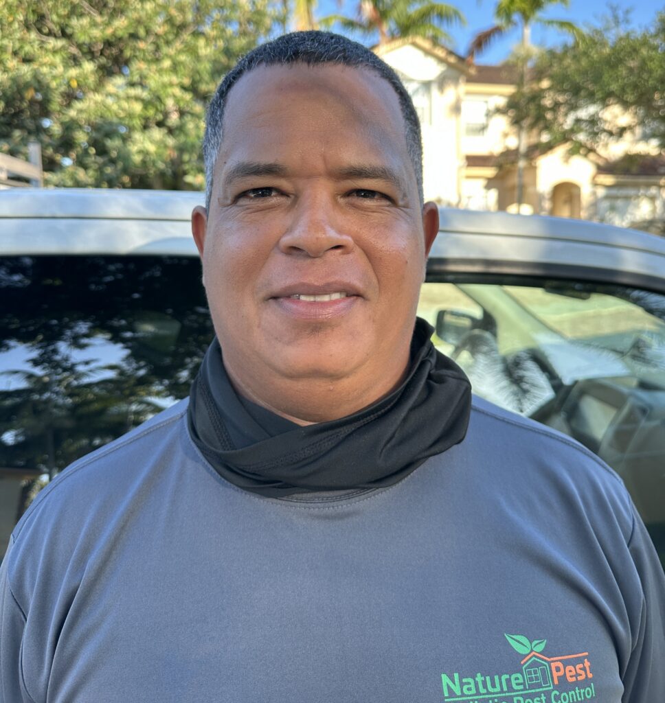 A man smiling outdoors, wearing a gray Nature Pest Control shirt and a black neck gaiter, standing in front of a vehicle with trees and a house in the background.