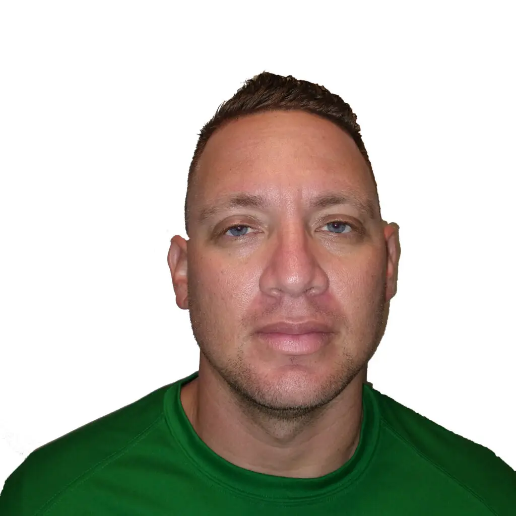 A man with short brown hair, light skin, and blue eyes is wearing a green crew-neck shirt, facing directly toward the camera with a neutral expression against a plain white background.