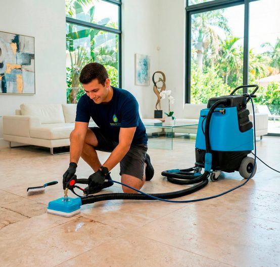 A man in navy blue work clothes performs tile and grout restoration with professional equipment in a bright, modern Miami living room featuring large windows and contemporary art.