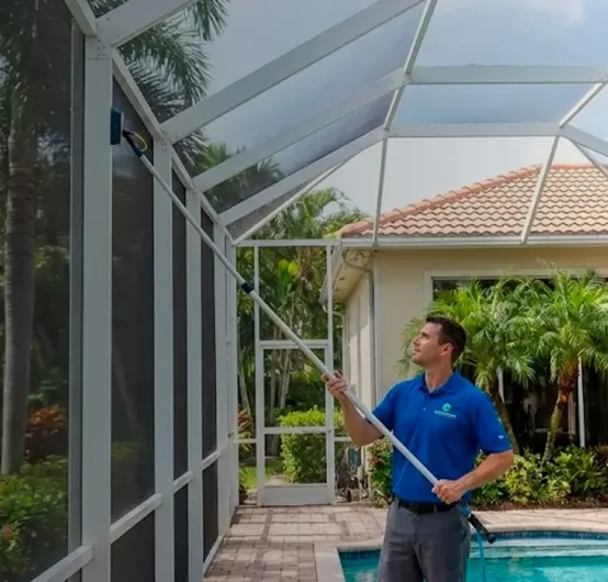A man in a blue shirt cleans the mesh screen of a backyard pool enclosure with a long pole brush. Palm trees and a beige house with a tiled roof are visible in the background.